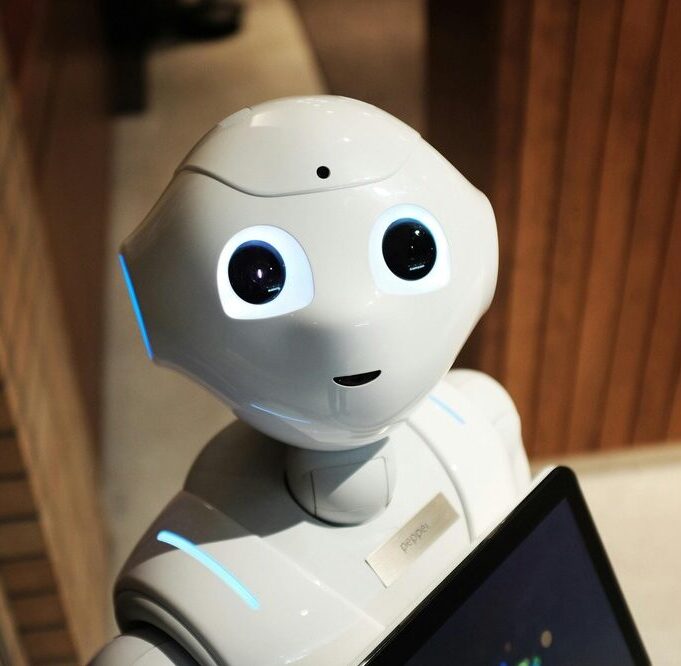 Close-up of a friendly white humanoid robot with large expressive eyes, holding a digital tablet in a modern indoor setting.