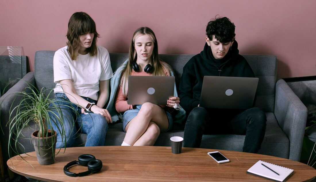 Three young individuals sitting on a sofa and collaborating on their laptops in a modern workspace environment.