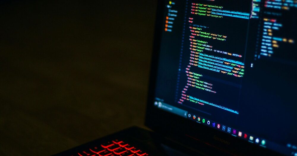 A laptop displaying colorful HTML/CSS code on the screen with a red backlit keyboard in a dark environment.