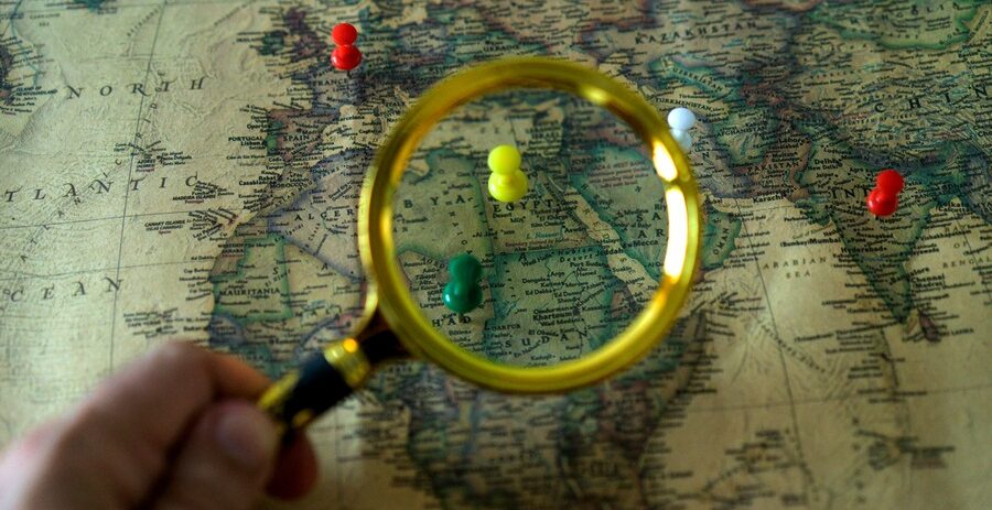 A hand holding a magnifying glass over a world map with various colored pushpins marking different countries.