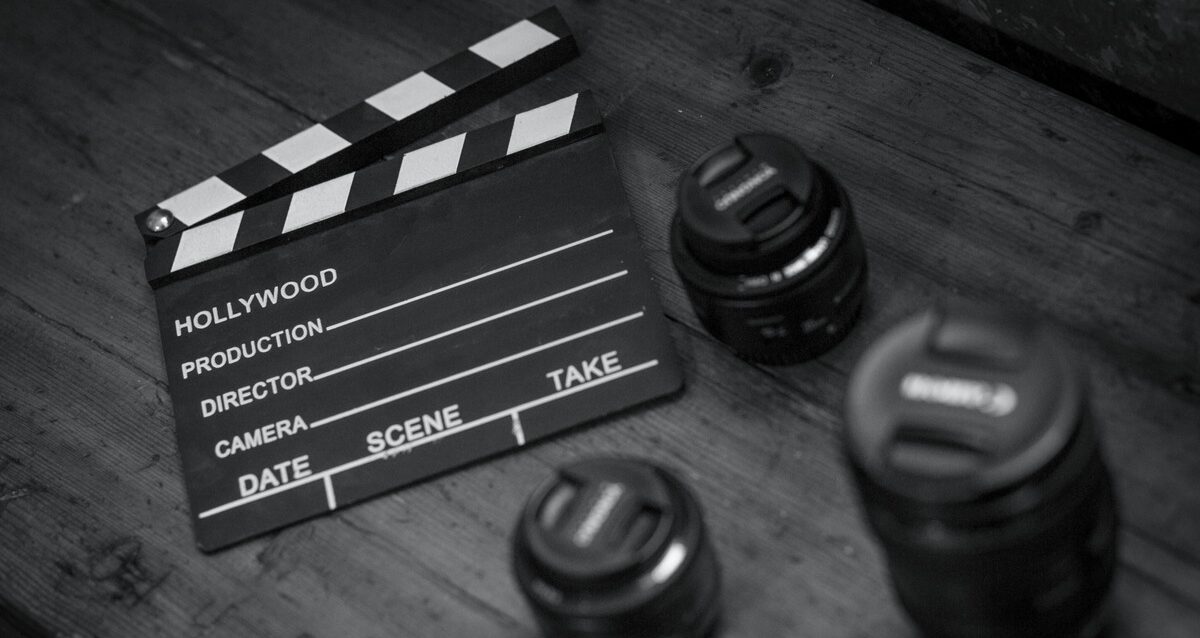 A black and white close-up shot of a Hollywood movie clapperboard lying on a wooden surface next to three professional camera lenses.