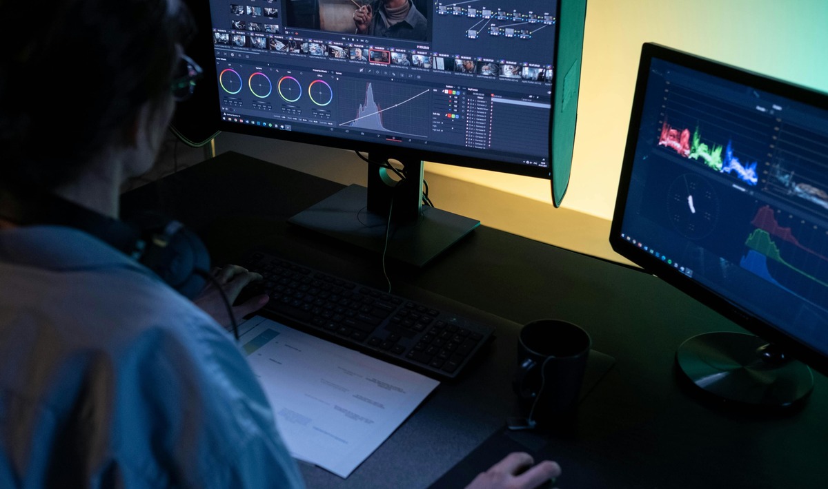 A professional video editor sitting at a dark desk, working on color grading and video post-production using multiple monitors with advanced editing software.