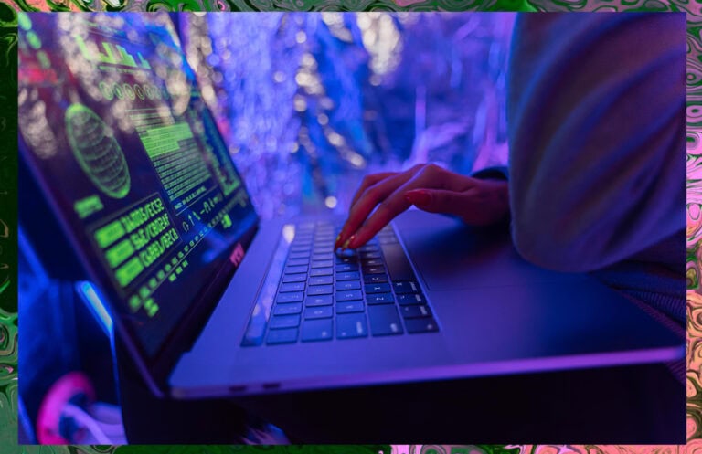 A close-up of hands typing on a laptop with green code and data graphs on the screen under neon blue and purple lighting.